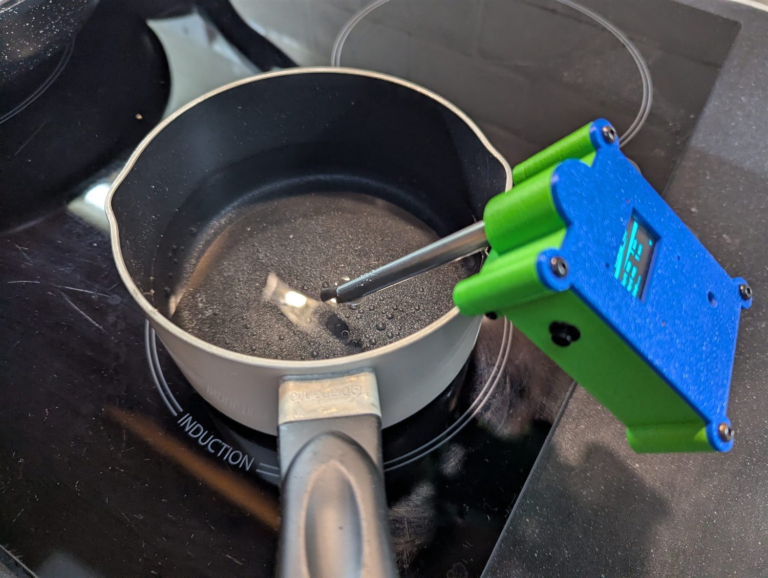 The device hanging on a saucepan on an induction hob, the word INDUCTION visible on the cooktop, display lit