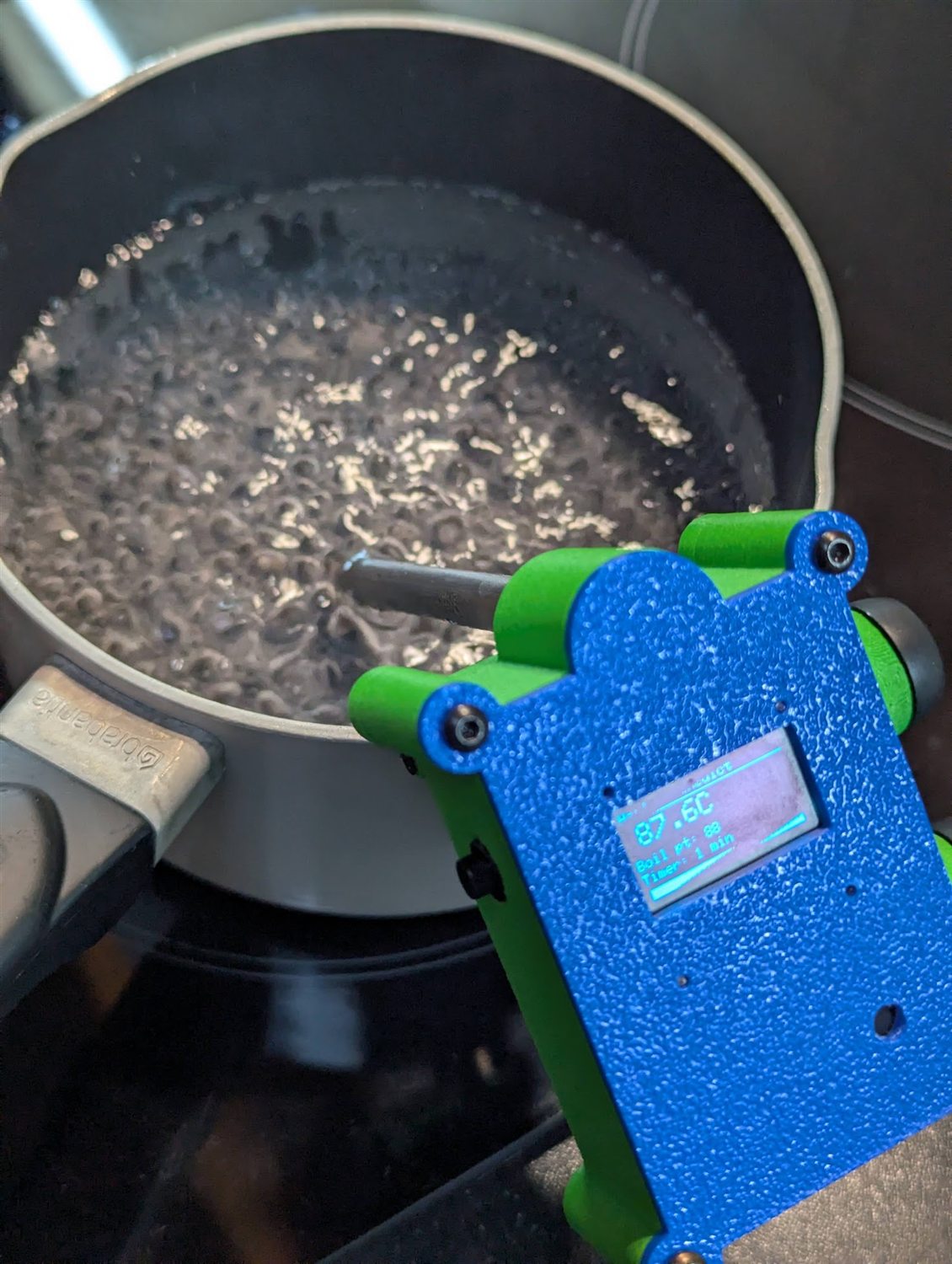 PotWatch clipped to a pot of heating water, its screen reading 87.6 degrees C with a boil point of 88 and a one-minute timer
